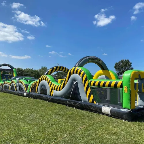 Long inflatable obstacle course with green and yellow details set up on a grassy field under a blue sky.
