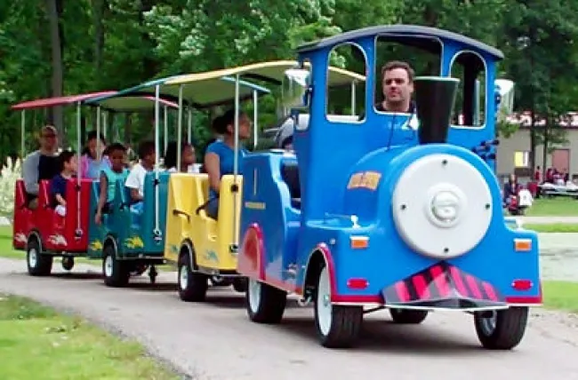 Colorful miniature train rides through park with adults and children enjoying a sunny day outdoors.