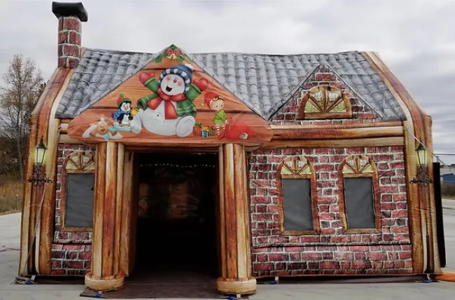 Inflatable house decorated with Christmas-themed snowman, elf, and penguin on a brick pattern exterior under gray sky.