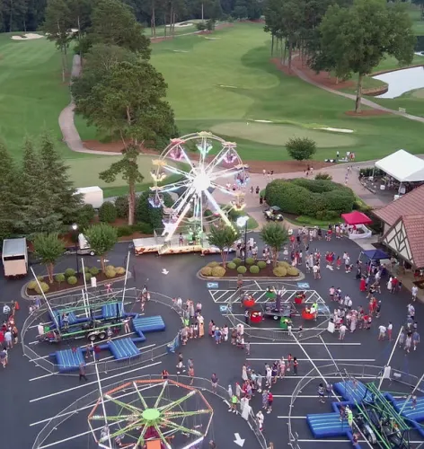 Evening view of a small carnival with illuminated Ferris wheels and rides near a golf course with green fairways.