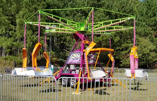 Colorful empty carnival swing ride with yellow, pink, and green accents surrounded by metal fencing on grass.