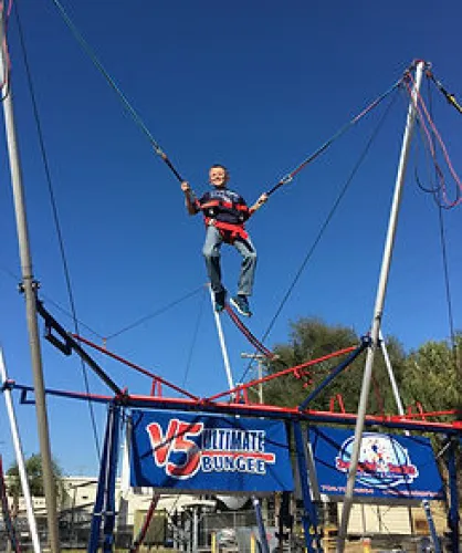 Child jumping on a bungee trampoline under clear blue sky at an outdoor amusement setup.
