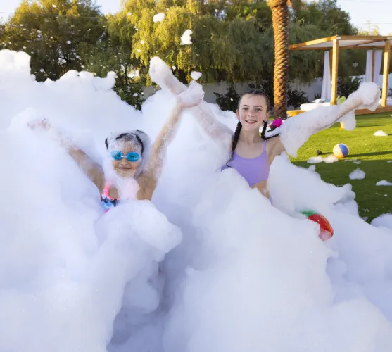 Two girls playing happily in a large foam party with foam covering their bodies and arms raised.