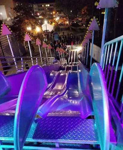 Nighttime view looking down a brightly lit multi-lane slide at a busy outdoor event with crowds and lights.
