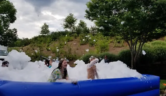 People enjoying a foam party in a blue inflatable pool outdoors surrounded by trees and plants.