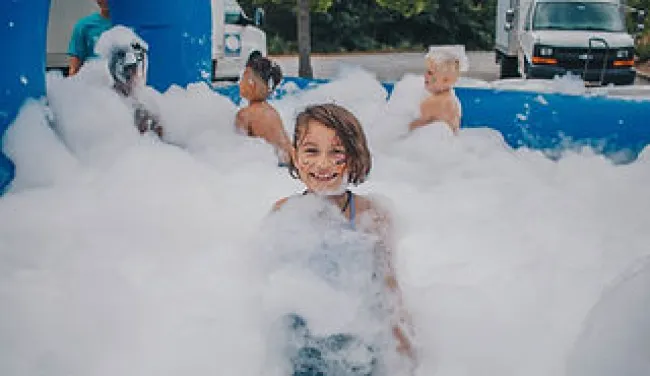 Children playing and smiling in a large foam pit outdoors during a sunny day.