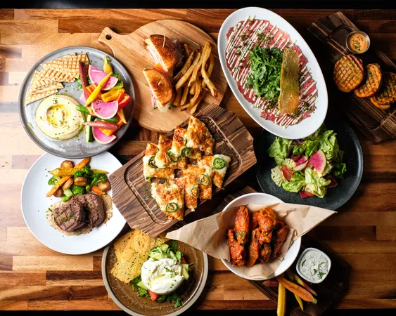 Overhead view of assorted dishes including flatbread, wings, salad, steak, sandwiches, and dips on a wooden table.