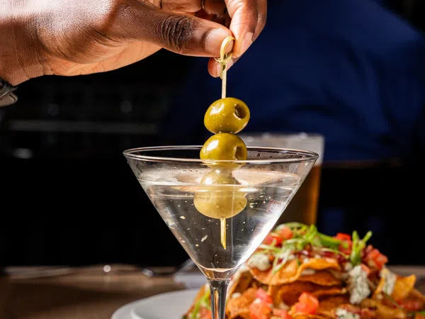 Hand holding skewer with green olives above a martini glass next to a plate of loaded nachos on a wooden table.