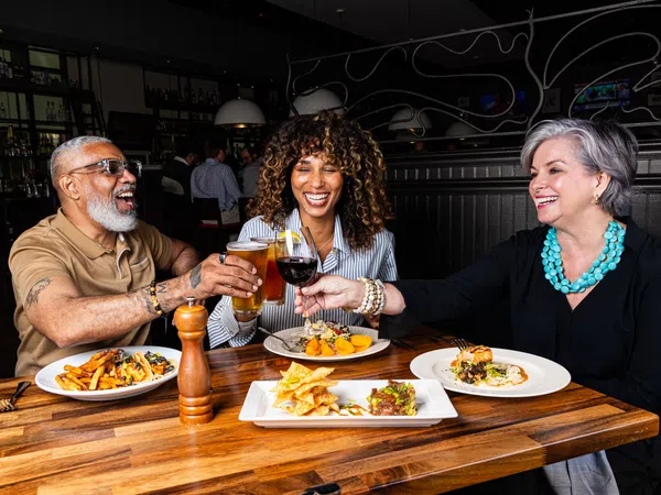 Three diverse adults enjoying drinks and gourmet food together at a wooden table in a restaurant.