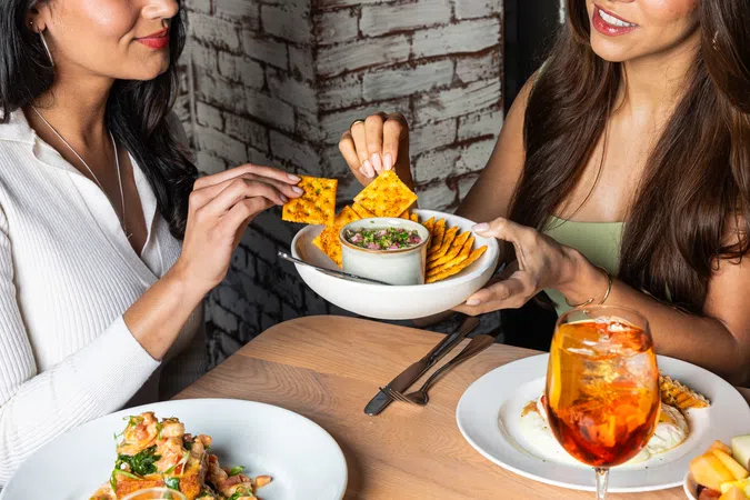 Two women sharing chips and dip while enjoying a meal with drinks at a cozy restaurant table.