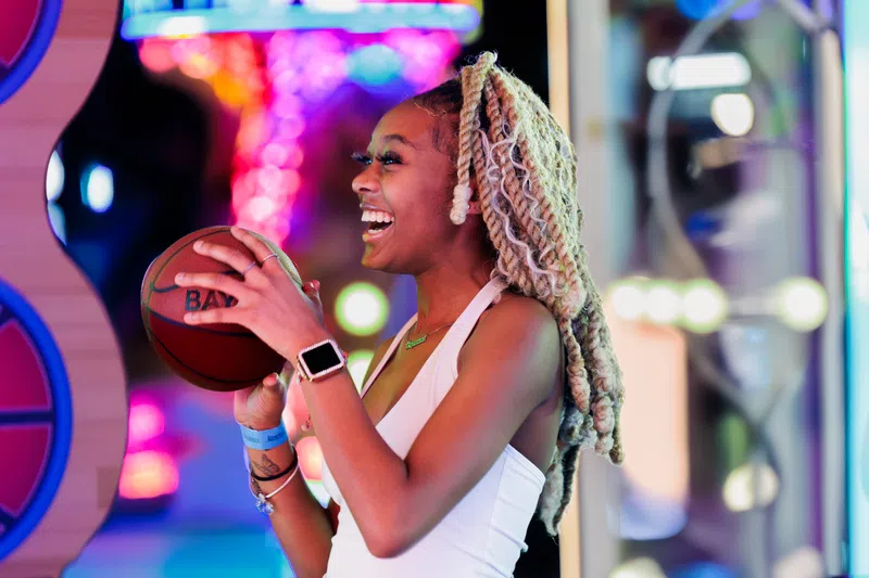 Young woman with blond twists laughing while holding a basketball at a vibrant, colorful arcade game.