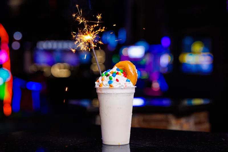 Creamy milkshake with whipped cream, colorful candy, donut, and lit sparkler in a dimly lit setting.