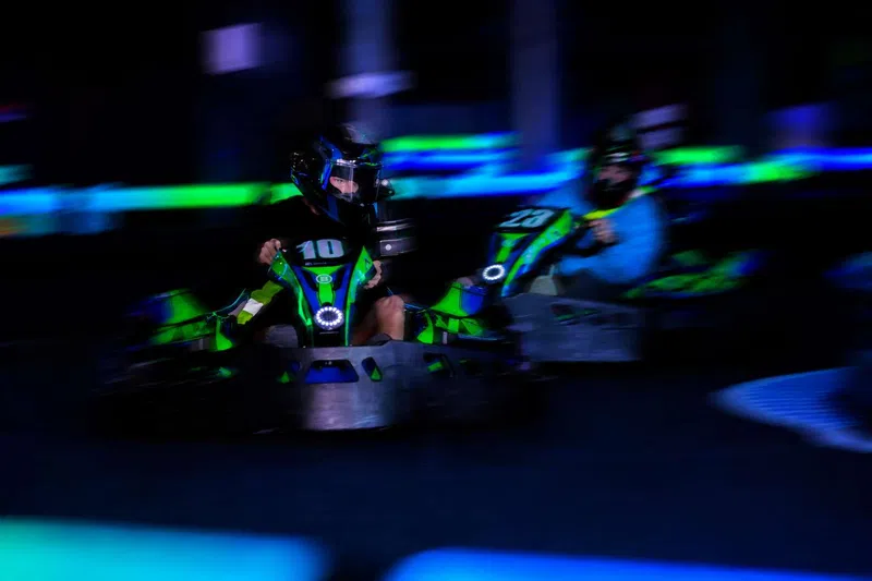 Two racers in green and black go-karts speeding on an indoor track with neon blue and green lighting.