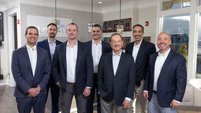 Group photo of diverse professionals in business attire inside an office with Reliant Real Estate branding.
