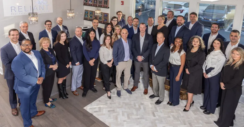 Group photo of diverse professionals in business attire inside an office with Reliant Real Estate branding.