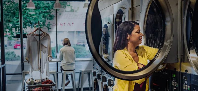 Woman loading clothes into front-load washing machine in cozy laundromat with large windows and seated customer.