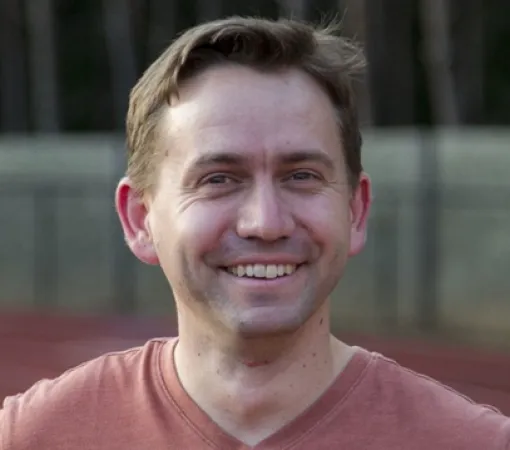 Smiling man with short brown hair wearing a rust-colored shirt outdoors on a blurred background.