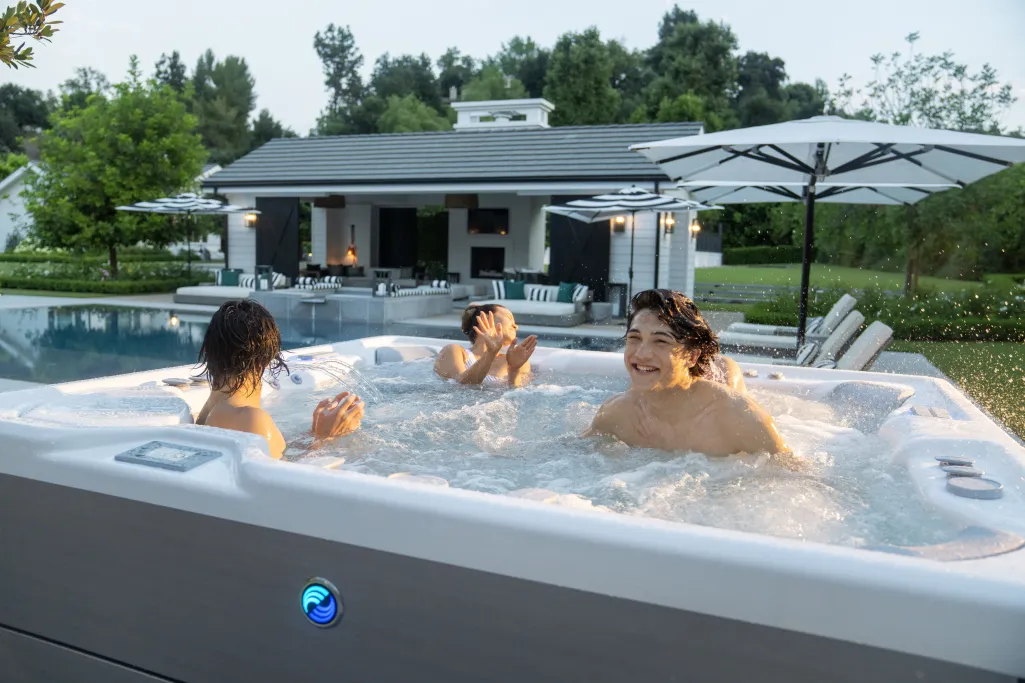 Three people enjoying a bubbling hot tub outdoors near a pool and patio with umbrellas at a modern house.