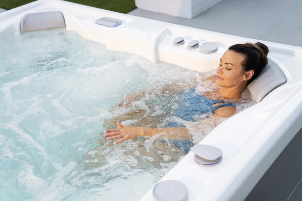 Woman relaxing in bubbly white hot tub outdoors with eyes closed and hands in water.