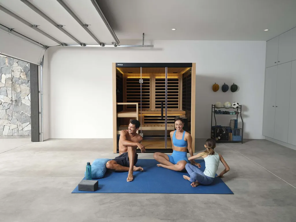 Family doing yoga on a blue mat in a modern garage with a glass sauna behind them