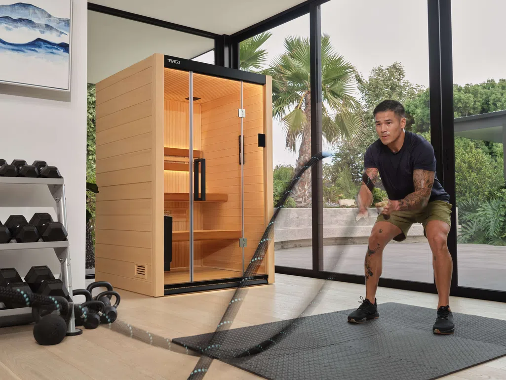 Man exercising with battle ropes next to a wooden infrared sauna in a bright home gym with large windows.