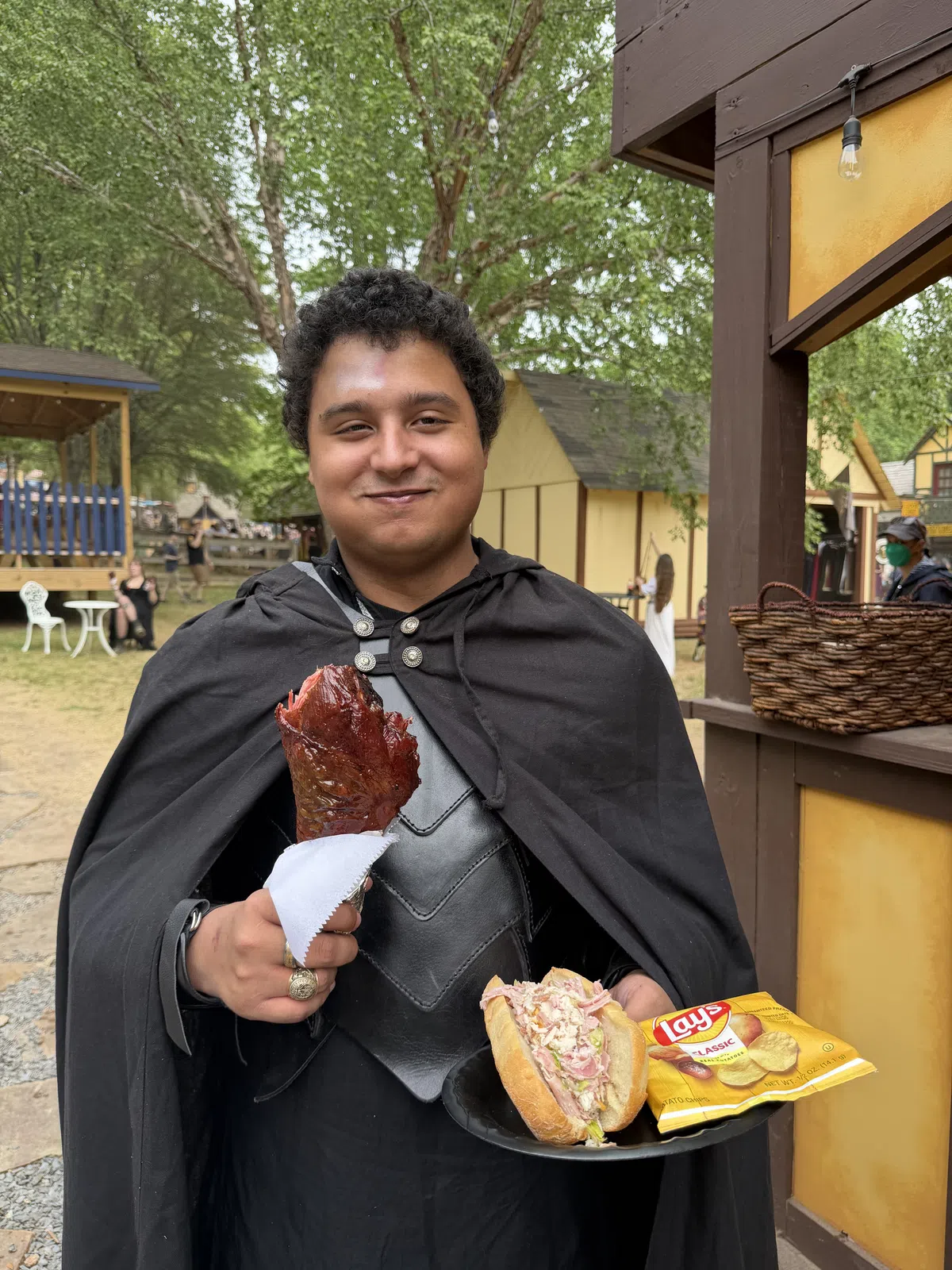 A guest in a black cape holds up a turkey leg, a hoagie and chips in hand