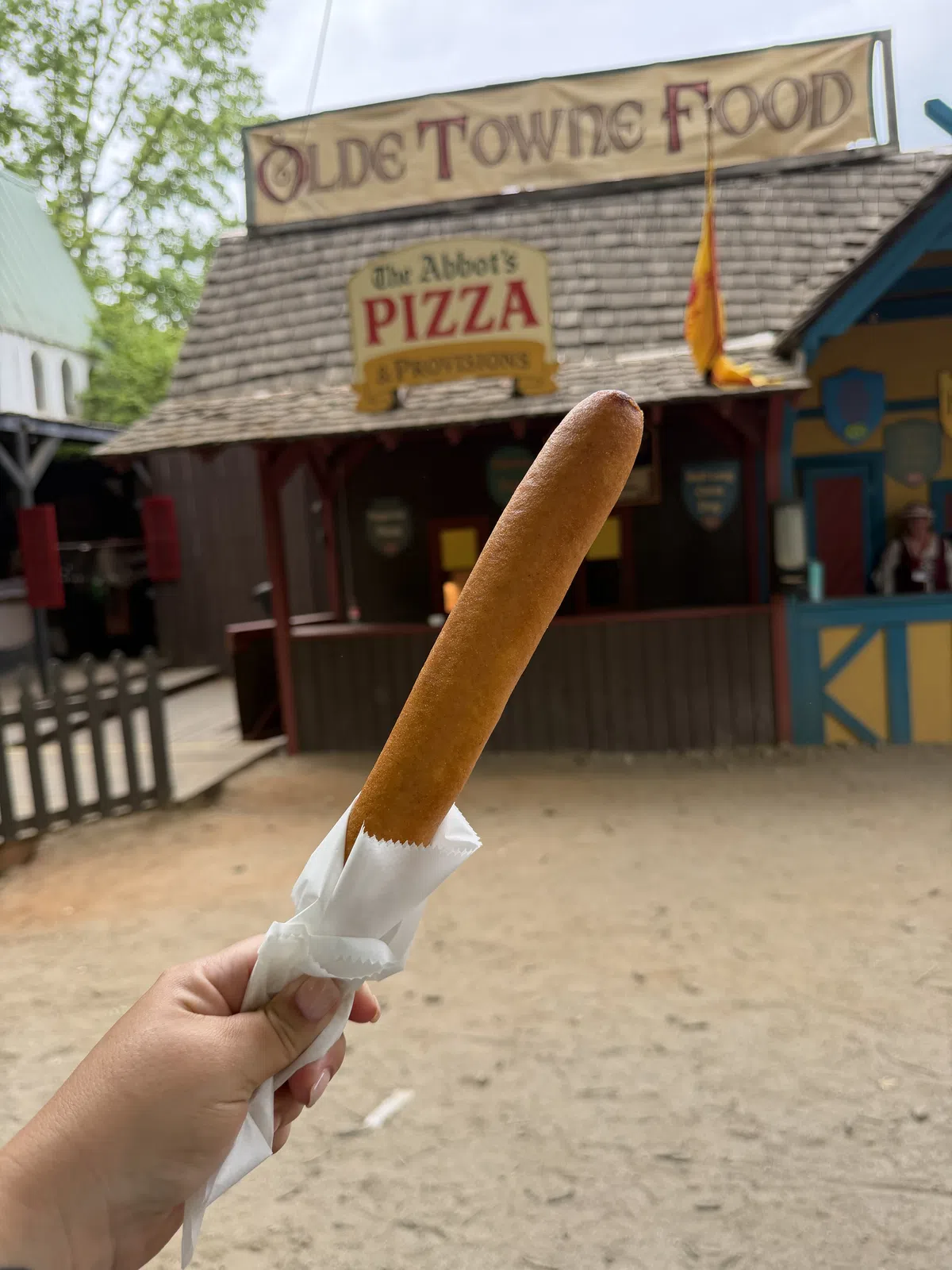 A foot-long corn dog held up before the Olde Towne Food banner and The Abbot's Pizza