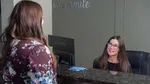 Receptionist smiling and assisting a woman at a clinic front desk with computer and sign behind.