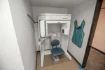 Dental X-ray machine with blue chair and protective lead apron hanging on wall in a clinical room.