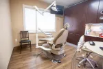 Modern dental office room with beige patient chair, equipment, wood cabinets, and hardwood flooring under bright light.