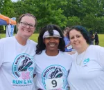 Three smiling women wearing matching 5K run event shirts posing outdoors by a finish line banner.