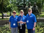 Three veterans in blue shirts and hats pose in front of the Vietnam Veterans Memorial statue in a sunny park.