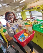 Smiling woman holding a shopping basket with groceries and a card inside a colorful fresh produce store.
