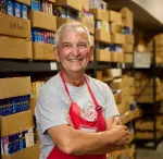 Smiling older man in red apron standing with arms crossed in a pantry filled with boxed food items.