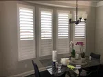 Dining room with four tall windows with white shutters, black table, orchids, and modern chandelier.