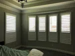 Room with five narrow windows featuring white plantation shutters and a green bedspread in dim lighting.