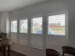 Four white plantation shutter windows in a bright room with minimal furniture and a view of houses outside.