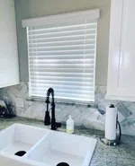 Modern kitchen sink with black faucet, white double basin, granite countertop, paper towel holder, and window blinds.