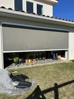Exterior view of a modern house with a large retractable screen partially lowered over a patio with plants.