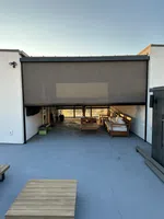 Modern outdoor patio with wooden furniture and retractable screen partially lowered on a smooth concrete floor.