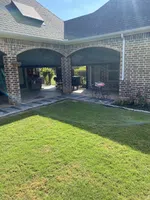 Sunlit backyard with green lawn, brick patio with arches, outdoor furniture, and a small decorative cart.