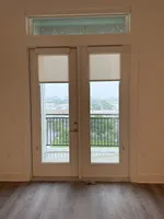 Double glass doors with beige roller shades open to a balcony overlooking a rainy cityscape with trees and buildings.