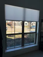 Window with a partially lowered white roller shade overlooking a suburban street with houses and cars outside.