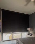 Bedroom with gray walls, black blackout window shades, bedside table, lamp, and a bed with gray bedding.