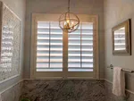 Modern bathroom with marble tiles, white shutters on window, hanging chandelier, towel rack, and framed wall art.