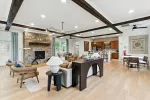 Spacious living room with exposed wood beams, stone fireplace, modern furniture, and open kitchen layout.