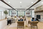 Spacious modern living room with stone fireplace, wooden beams, comfortable seating, and large windows with curtains