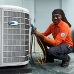 Technician in orange uniform servicing Carrier Infinity HVAC unit with gauge and hoses, smiling at camera.