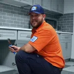 Plumber wearing an orange shirt and blue cap repairing kitchen plumbing under the sink with a wrench.