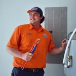 Smiling plumber in orange Estes uniform holding red pipe wrench near appliance and electrical panel